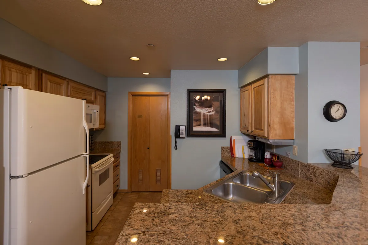 A cozy kitchen view focusing on a granite countertop, modern kitchen appliances, framed artwork, and a wall clock. The intimate lighting and layout invite families or groups to gather and create meals together, fostering a sense of togetherness and comfort.