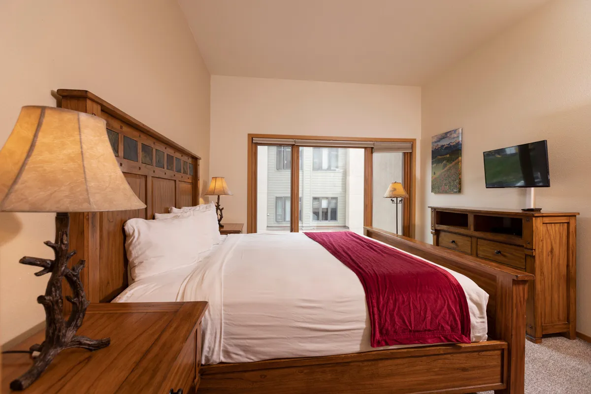 A king-size bed with white linens and a red throw blanket is complemented by rustic wooden furniture. Large windows let in natural light, providing serene mountain views and creating a tranquil retreat.