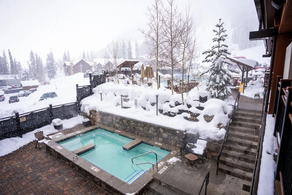 An inviting rooftop hot tub nestled amidst a snow-covered landscape, framed by surrounding trees and a staircase, with steam rising from the water and a serene wintry backdrop.