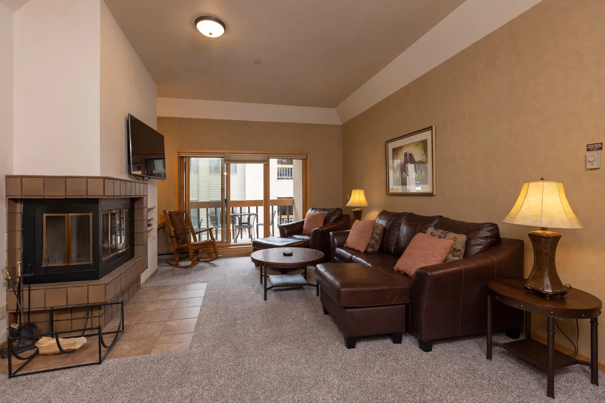 The image showcases a cozy condo living area with a crackling fireplace, inviting sofa, and a coffee table set for relaxation. Natural light pours in through a sliding glass door that leads to a balcony, revealing glimpses of the surrounding mountains. The scene evokes warmth, comfort, and the perfect après-ski setting.