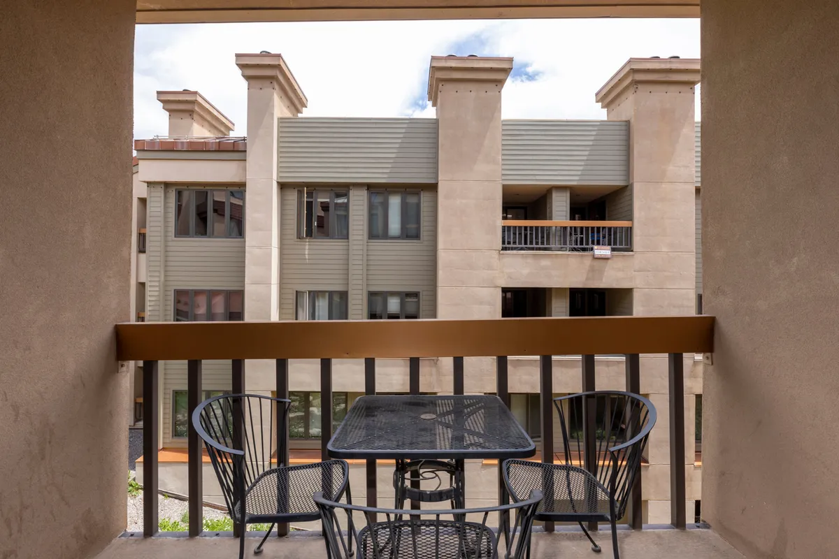 A bright exterior shot featuring a balcony with a table and chairs, set against a backdrop of blue skies and adjacent buildings. The image evokes a sense of openness and possibility, inviting guests to start their day with sunrise views or unwind while stargazing or watching the sunset.