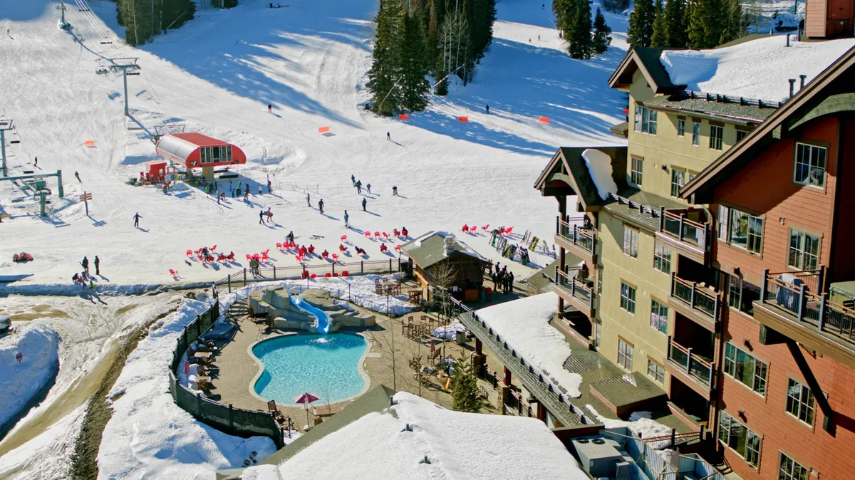 A dynamic view of the ski resort with sunlit ski slopes, skiers in action, a resort swimming pool, and the lodge. The architecture blends with the snowy landscape, highlighting both the excitement of winter sports and relaxation options like the pool and social spaces.