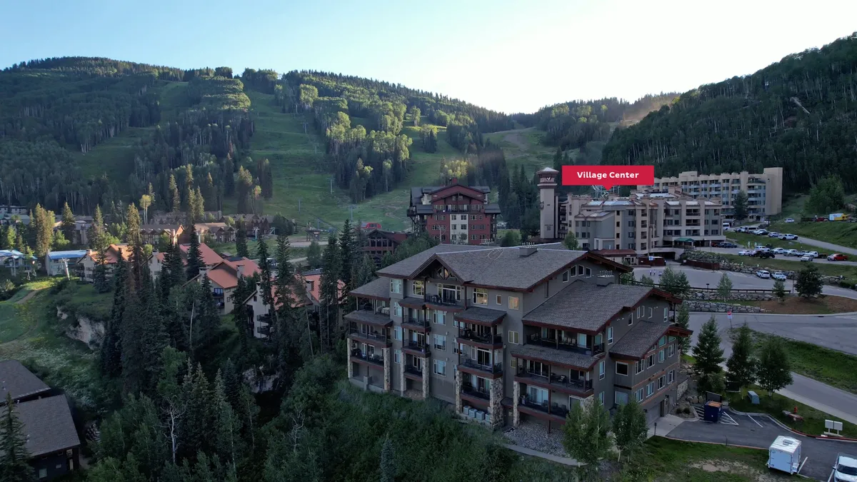 An inviting view of the resort village nestled among mountains and forests, with hints of parking and pathways leading to adventure. The unique architecture and lively feel suggest a hub of activity year-round.