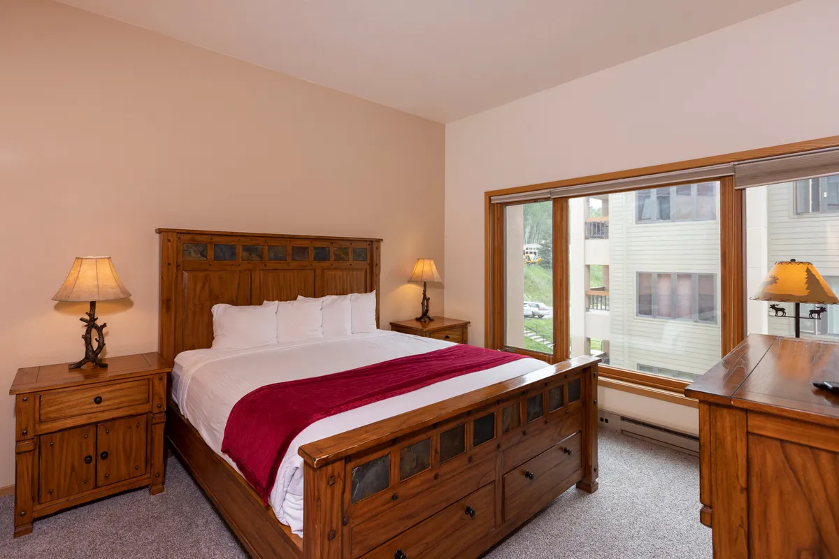 A cozy bedroom featuring a wooden bed adorned with a red throw blanket, softly glowing bedside lamps, and ample natural light filtering in. The space conveys warmth and comfort, ideal for a restful night's sleep.