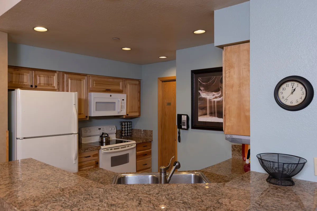 A warmly lit kitchen scene highlighting a granite countertop, modern appliances, wooden cabinetry, framed wall art, and a wall clock. The view extends to the spacious counters and functional appliances, suggesting an environment suitable for meal prep and group cooking. The inviting setup encourages family gatherings and shared kitchen experiences.