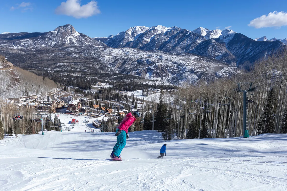 A lively winter scene on the slopes, featuring a skier and a child enjoying the snow with majestic mountains in the background. The ski resort buildings and lifts are visible, framing a picture-perfect family adventure.