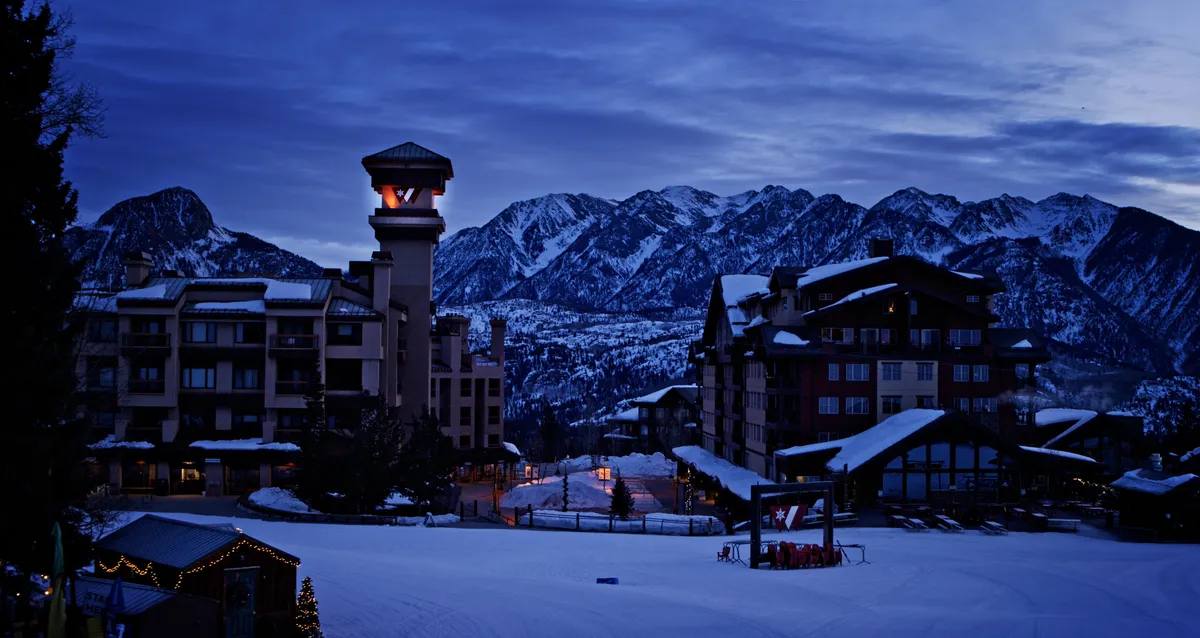 A picturesque winter evening scene with snow-capped mountains glowing at sunset, warmly lit resort buildings, and festive winter decorations. The ambiance is peaceful yet inviting, evoking the cozy, luxurious side of the adventure.