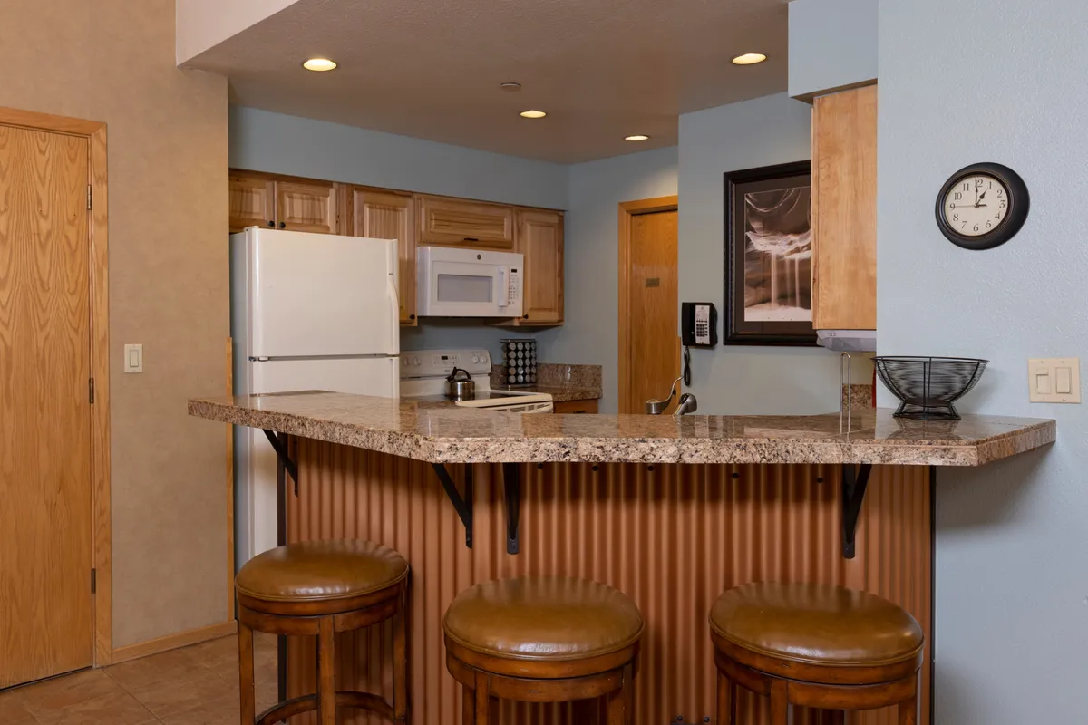 A bright, open kitchen featuring granite countertops, wooden cabinets, bar stools at the counter, a prominent wall clock, and framed artwork. The space feels warm and inviting, with plenty of room for multiple people to gather or cook together. The open counter with stools encourages casual conversation and socializing while meals are being prepared.