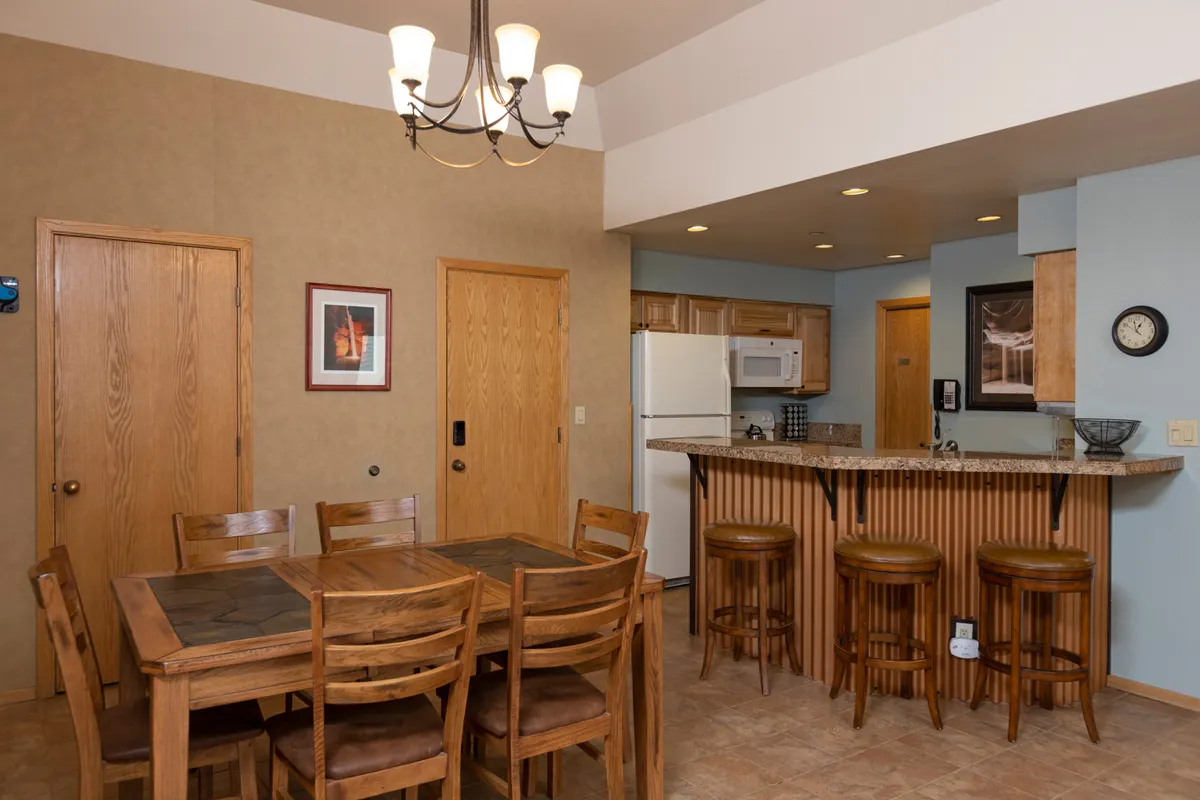 The dining area is shown with a central dining table, a kitchen bar, and a stylish chandelier above. The setup suggests a communal space for meals, entertaining guests, and enjoying après-ski snacks or games.